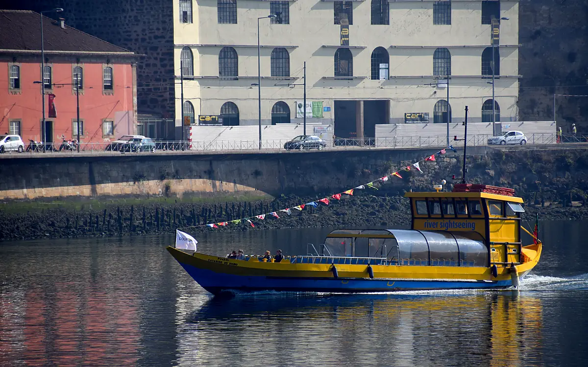 De geel-blauwe veerpont met vlaggetjes vaart over de Douro langs de kade, met een gebouw met witte kozijnen aan de overkant