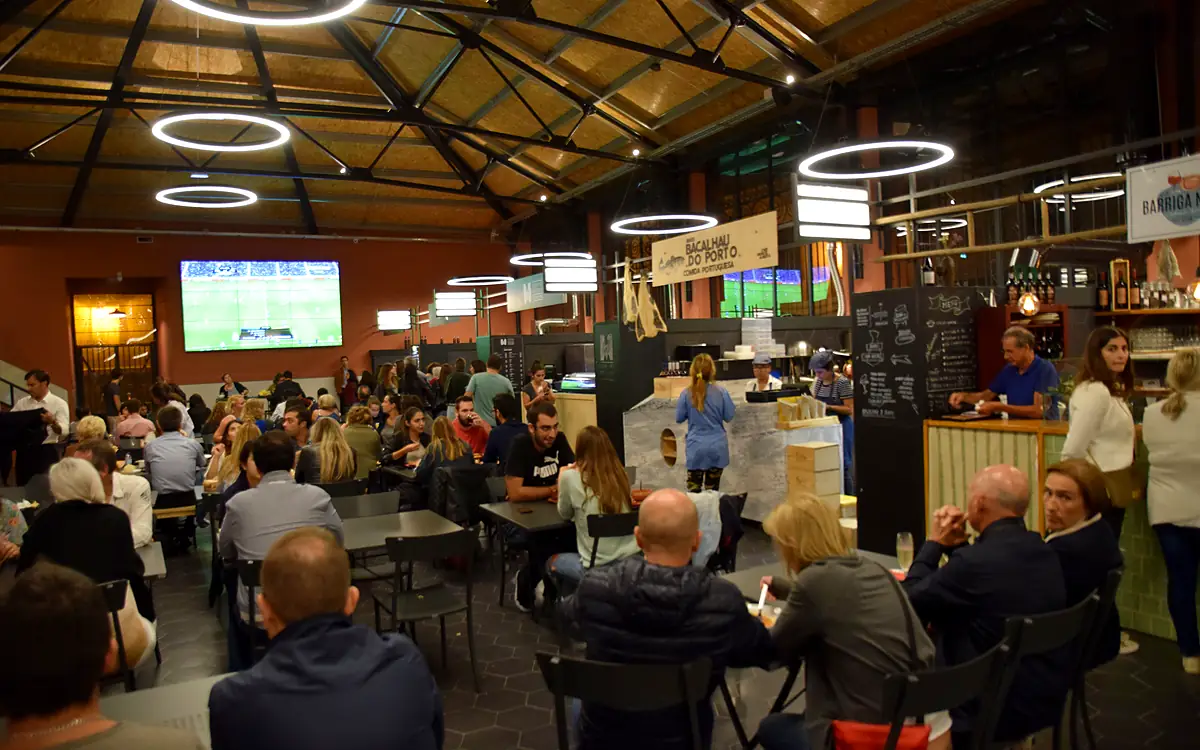 Bezoekers aan lange tafels onder ringlampen in een overdekte markthal