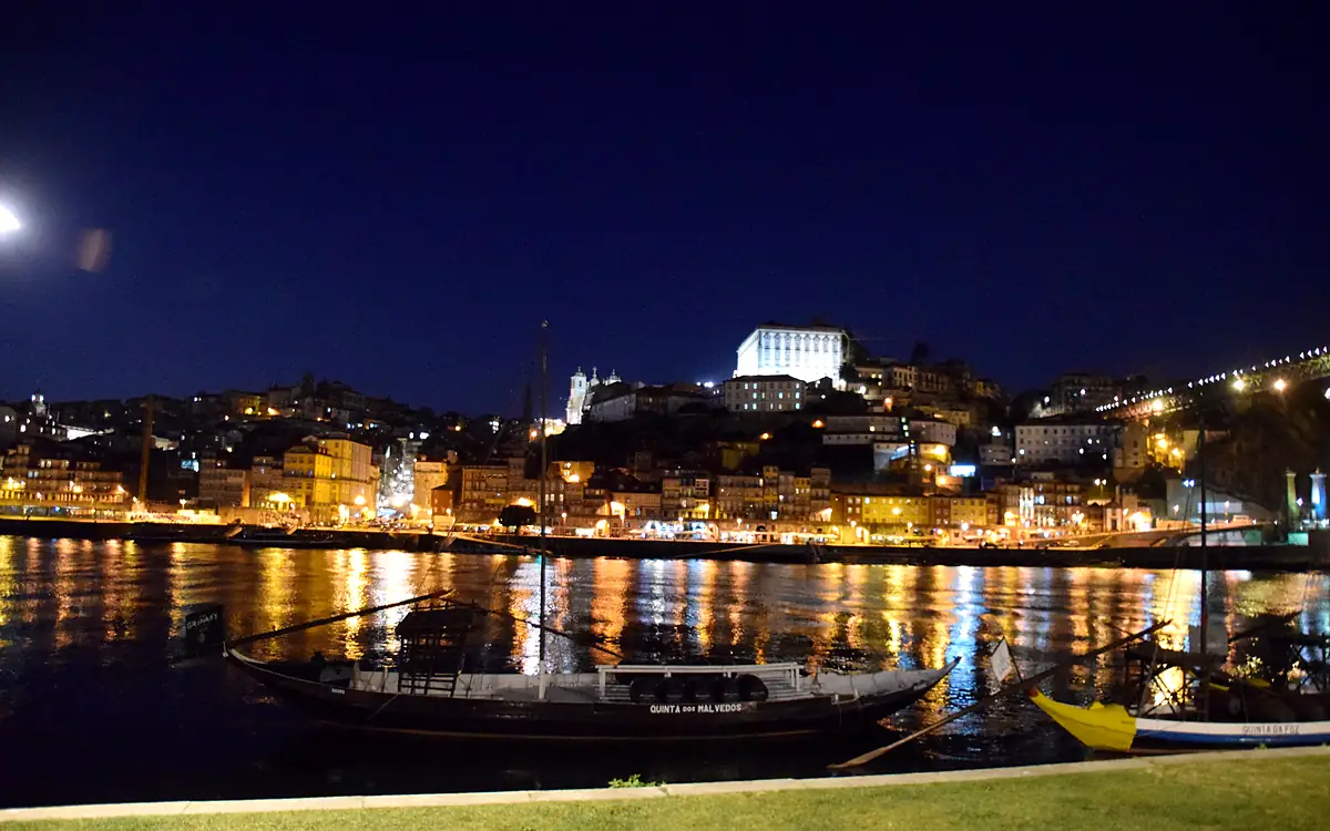 Porto bij nacht vanaf de overkant van de Douro, met een rabelo-boot op de voorgrond