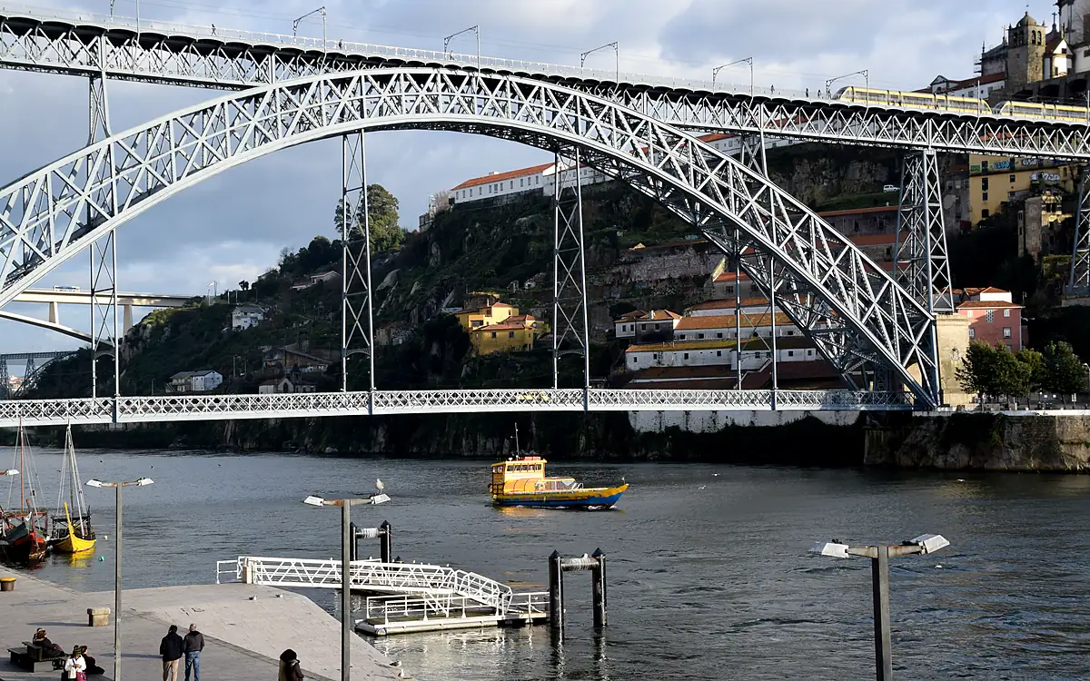 De Ponte Dom Luís I in volle breedte vanaf de kade, met de gekleurde huizen van Vila Nova de Gaia en een gele veerboot op de Douro